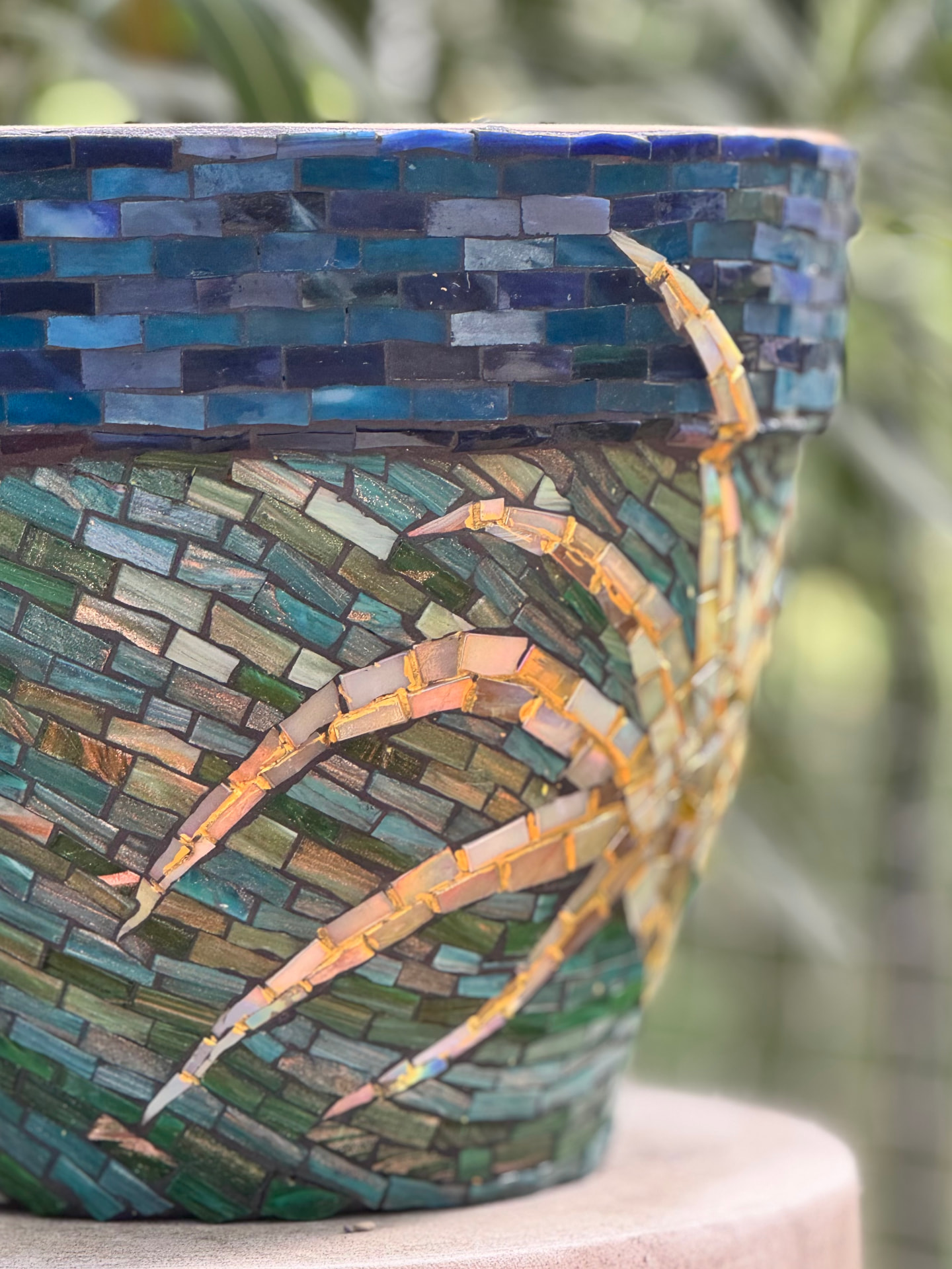 Handgefertigter Mosaik-Blumentopf in Türkis, Petrol und Grüntönen mit goldenen Kintsugi-Adern, vor grünem Gartenhintergrund.