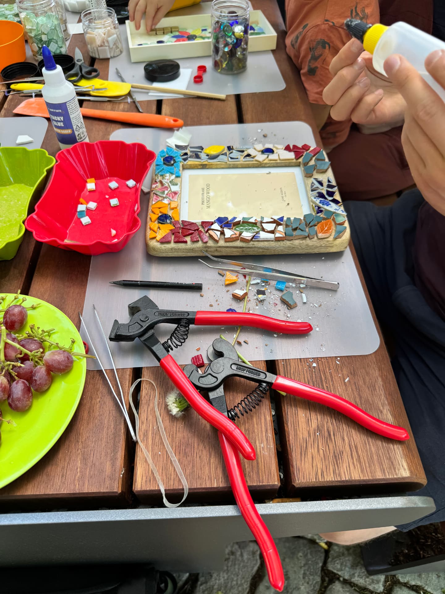 Werktisch im Mosaikworkshop im Freien mit halbfertigem Mosaik-Bilderrahmen, Zangen, Glassteinen und Kleber.