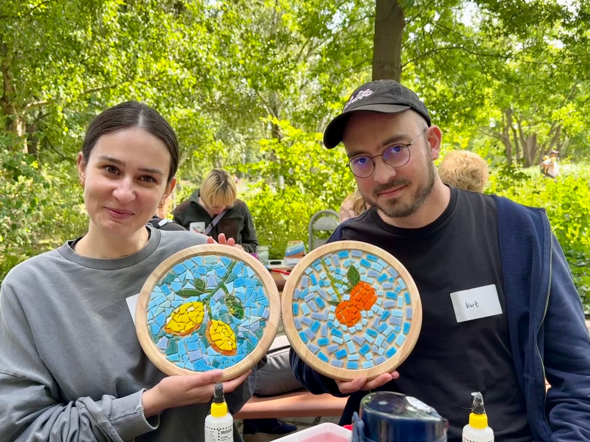 Zwei Teilnehmende zeigen ihre runden Mosaik-Werkstücke mit Zitronen- und Orangenmotiv
